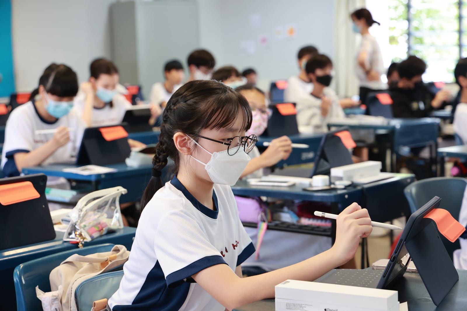 Students wearing face masks sit at desks using tablets with styluses in a classroom.